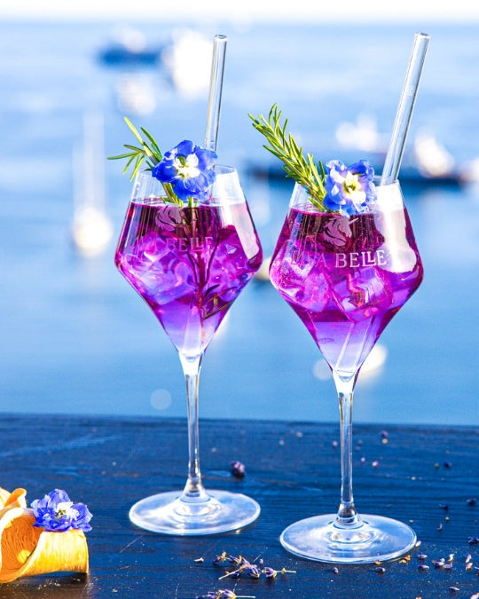 Zwei Lav'a Belle Signature Apéritif-Gläser mit graviertem Logo, gefüllt mit violettem Spritz vor Meerkulisse